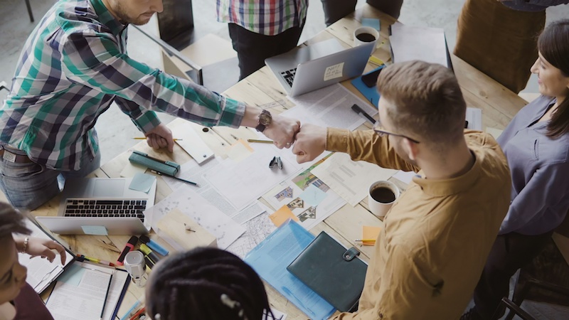 Top view of young business team working together near the table, brainstorming. Two mans fist greeting each other. Mixed race group of hipster people working at loft office on architectural design.