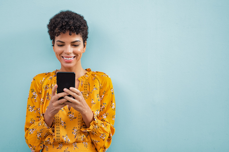 Happy smiling woman using smart phone. Young brazilian woman writing a message with mobile isolated on blue background. Portrait of african american stylish girl using cellphone with copy space.