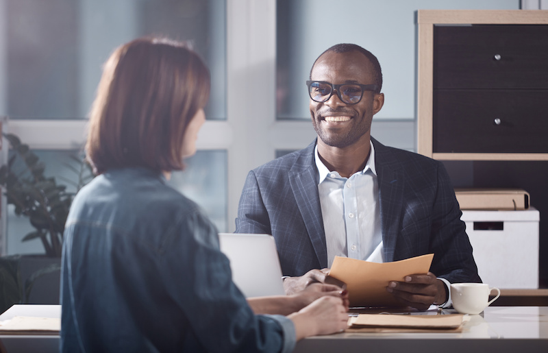 Portrait of optimistic young qualified manager is looking at his colleague female with smile while sitting at table and having pleasant communication. Back view of woman