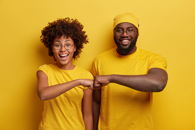 Studio shot of overjoyed satisfied woman and man, give fist bump, agree to work together, have success in project work, wear yellow attire, pose in studio. Cooperation concept