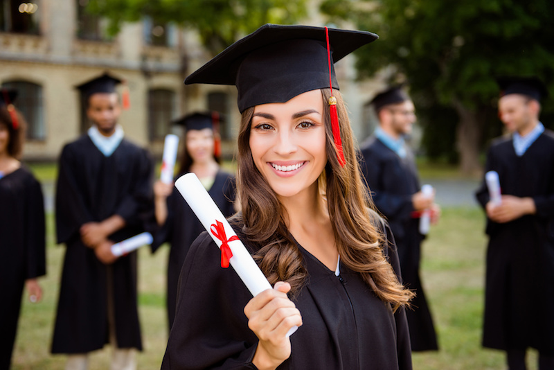 Happy brunette grad girl is smiling, blurred class mates are behind. She is in a black mortar board, with red tassel, in gown, with nice brown curly hair, diploma in hand