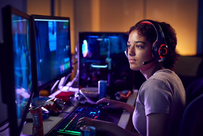 Girl Wearing Headset Gaming At Home Using Dual Computer Screens