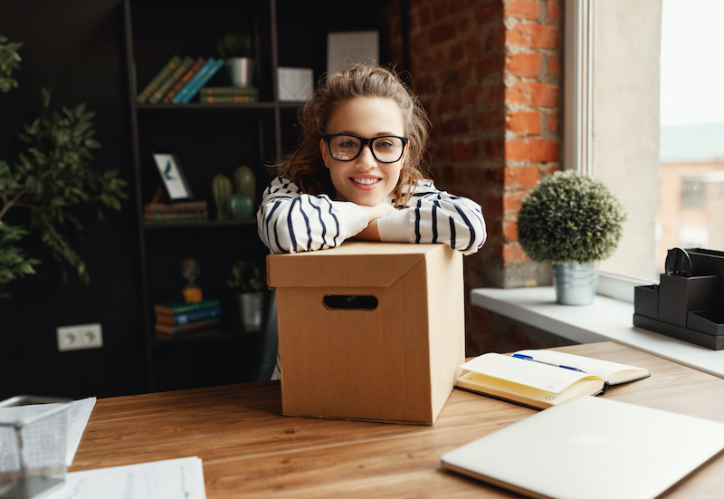 Young female newcomer in glasses and casual clothes smiling at camera while sitting at wooden table and leaning on carton box in dark modern office