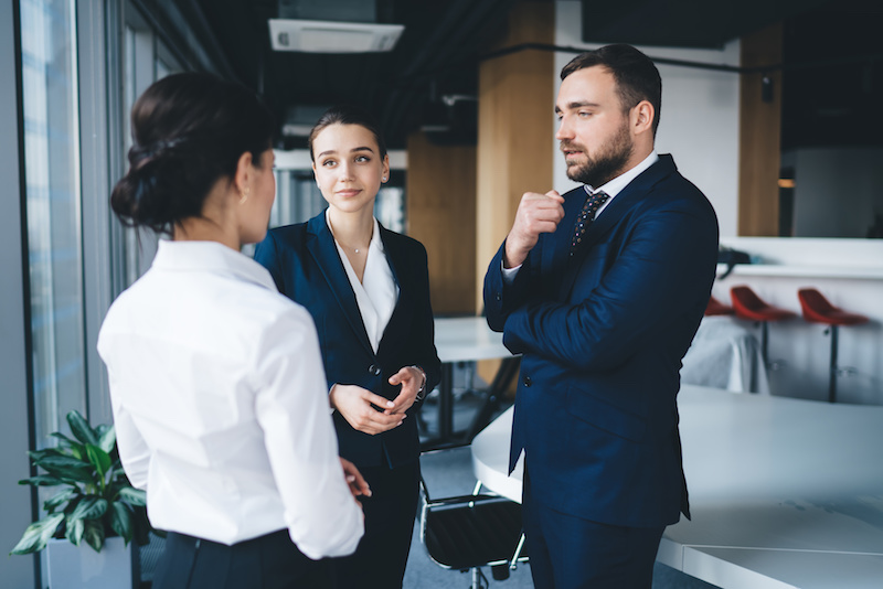 Lawyer coworkers in formal outfit talking and gesturing while standing together beside table at urban conference room in daylight